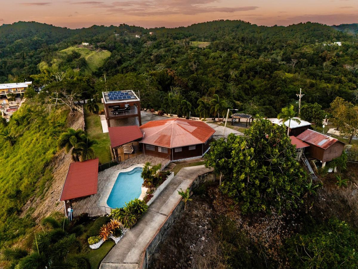 Vista aérea al atardecer de la hacienda de madera y su piscina, solas en la cima de una colina verde
